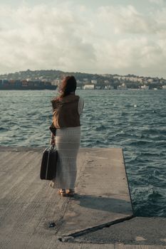 A woman stands on a pier with a suitcase, gazing at the sea and cityscape in the distance.