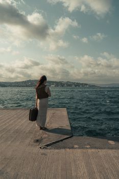 A woman with long hair stands on a concrete pier in İstanbul overlooking the sea with a suitcase.
