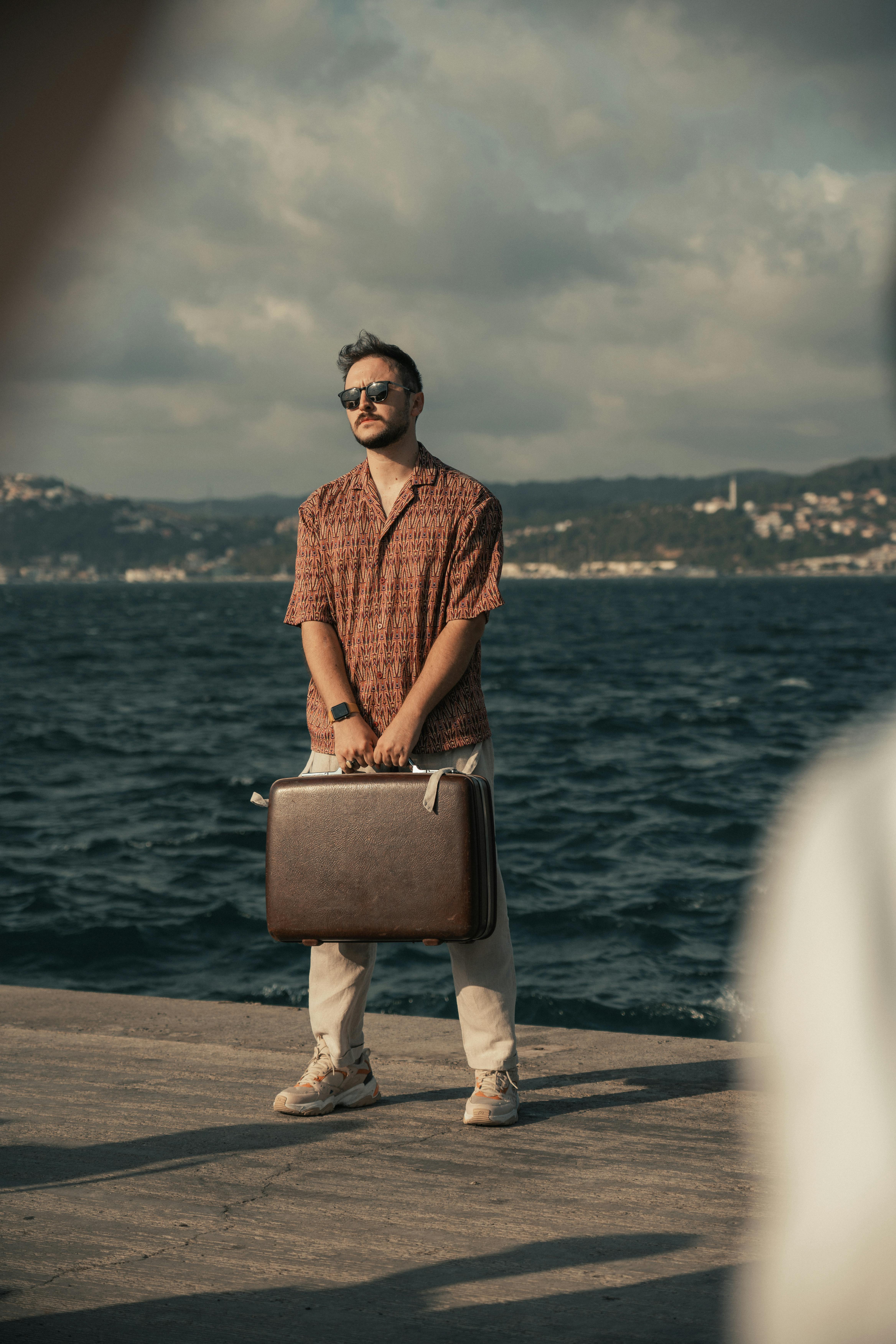 Stylish man in sunglasses holding a suitcase on a pier by the sea