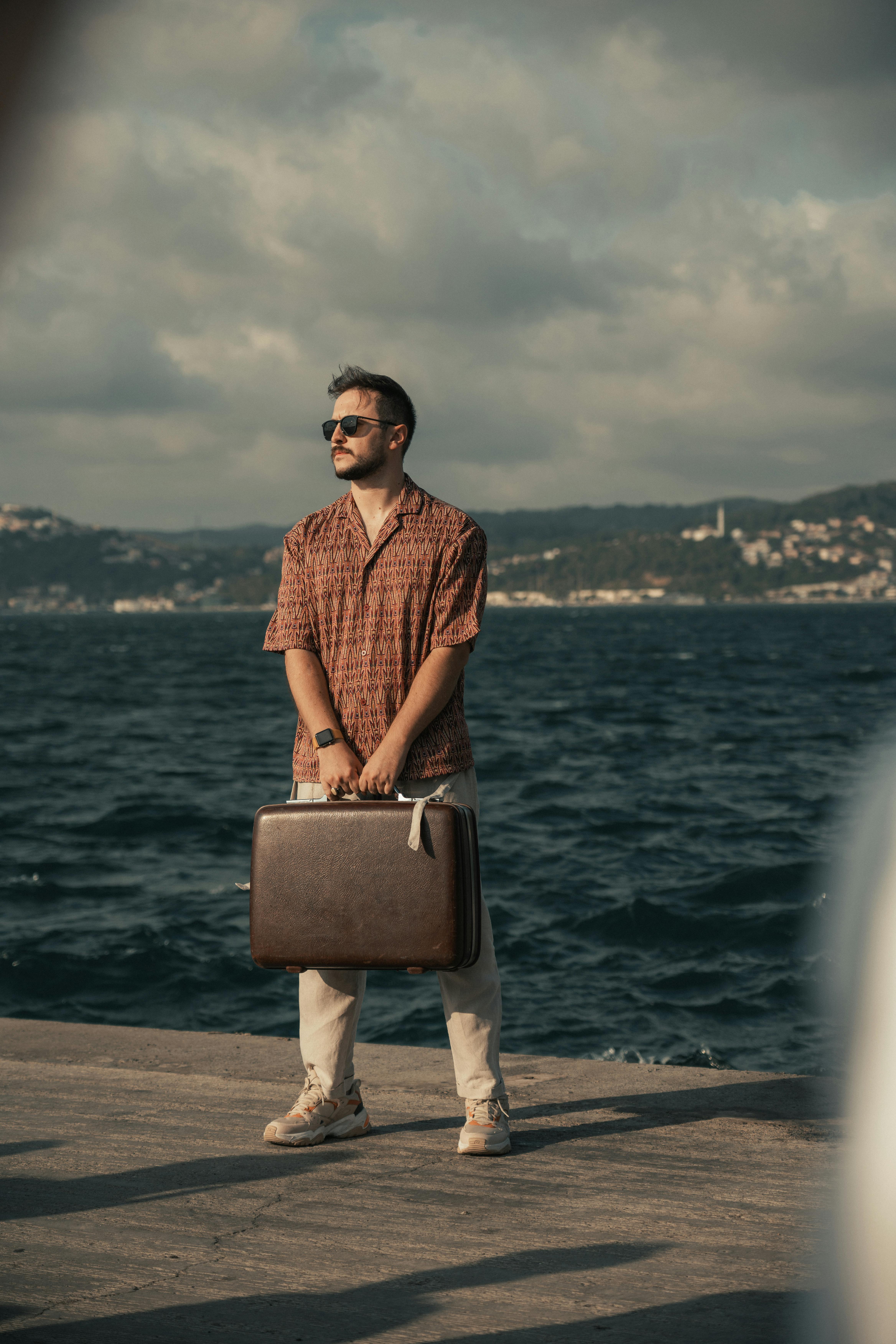 A man in a casual outfit holding a suitcase stands on a pier by the sea, facing the horizon.