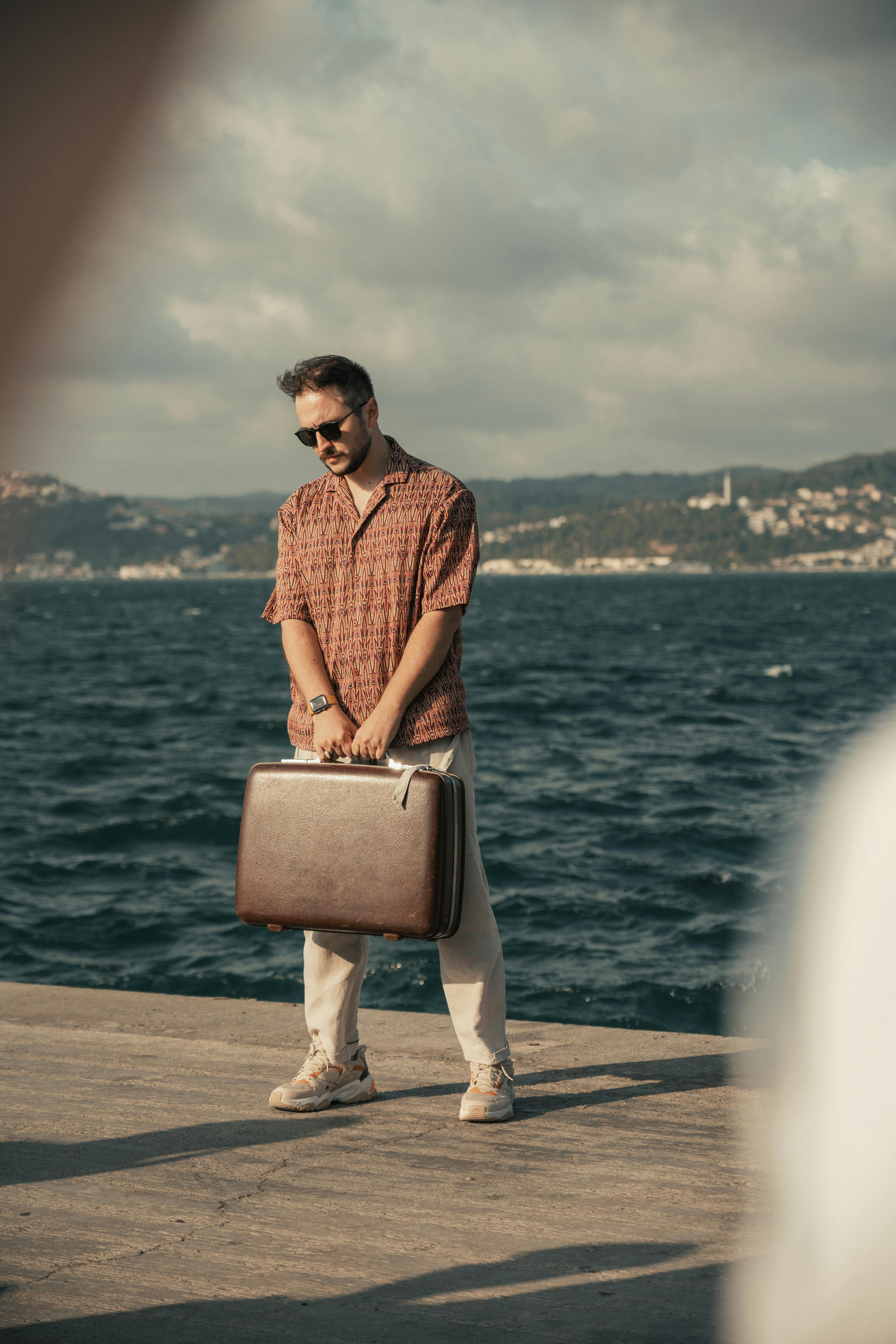 Stylish man holding a suitcase by the sea, wearing sunglasses and casual shirt.