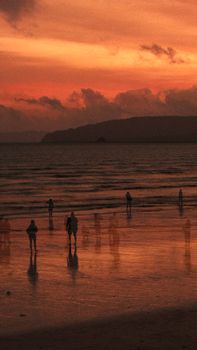 A vibrant sunset casts silhouettes of people standing on the shore, creating a dramatic beach scene.
