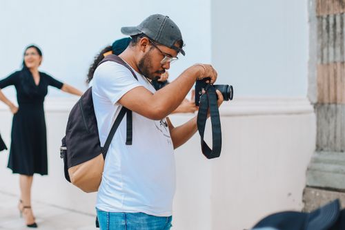Free A man in a white t-shirt and cap taking photos on a city street, focusing intently. Stock Photo