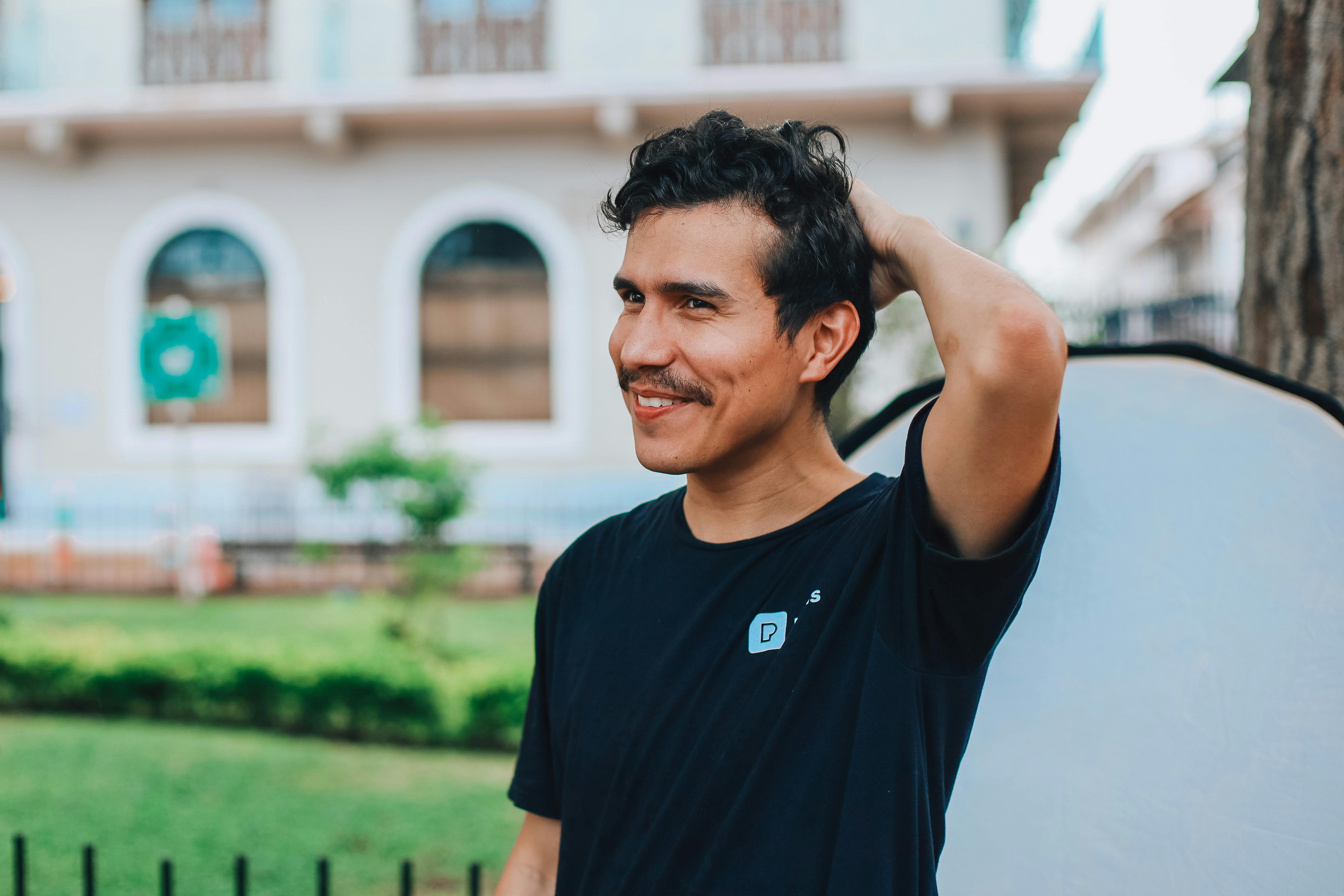A young man in a black t-shirt smiling outdoors, casually fixing his hair.