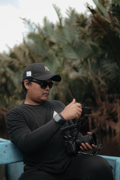 Free Man in black attire and cap holding camera while seated outdoors. Stock Photo