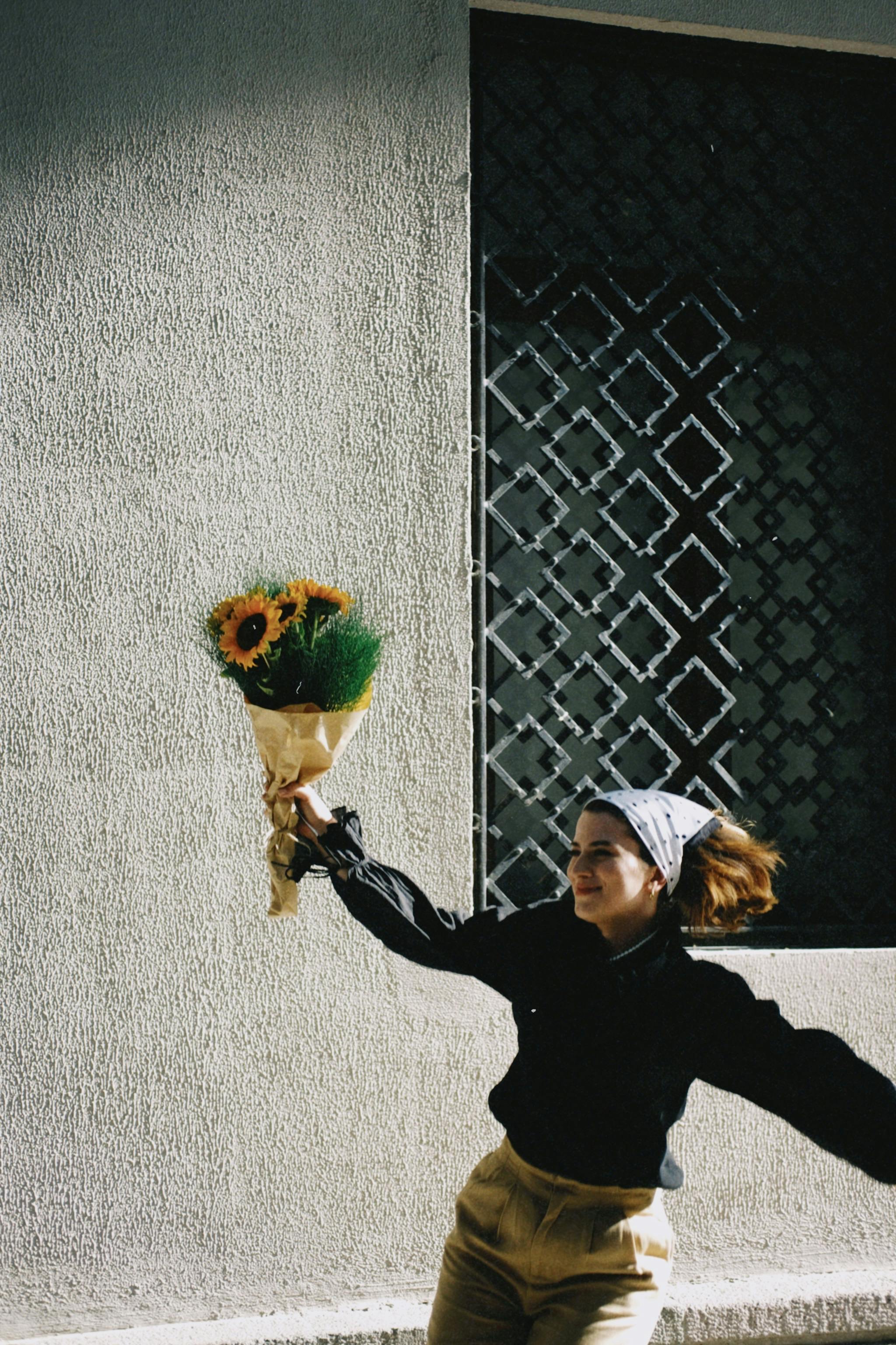 A woman with a cheerful expression holds a sunflower bouquet against a textured wall, enjoying a sunny day.