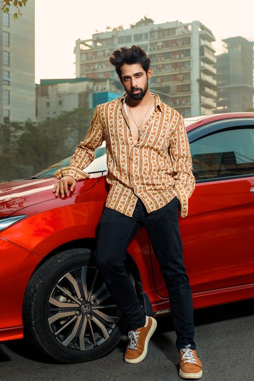 Handsome young bearded Indian man in a colorful shirt and jeans posing on the street near a red car