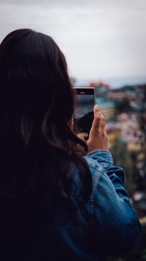Free A woman in a denim jacket photographs a cityscape using her smartphone from a vantage point. Stock Photo
