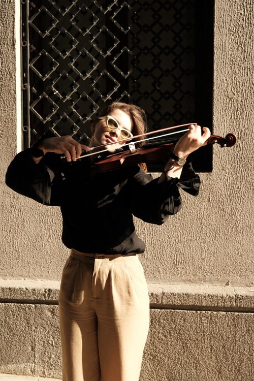 Free Woman in sunglasses plays violin outdoors in the sunlight, against a textured wall. Stock Photo