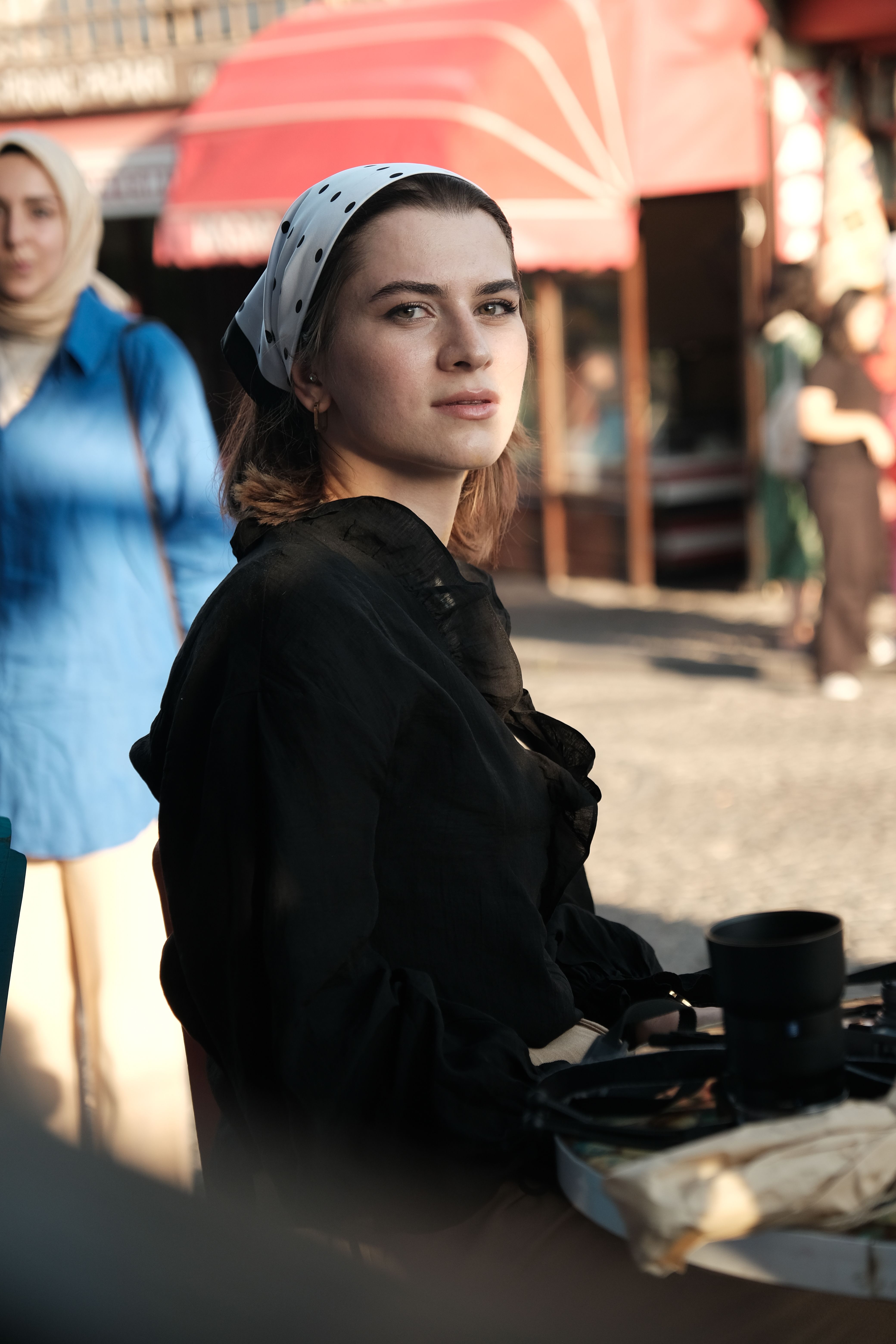 A young woman wearing a headscarf sits at an outdoor cafe, capturing a candid urban moment.