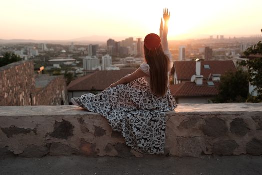 A young woman in a patterned dress enjoys a vibrant sunset from a city rooftop, embracing tranquility.