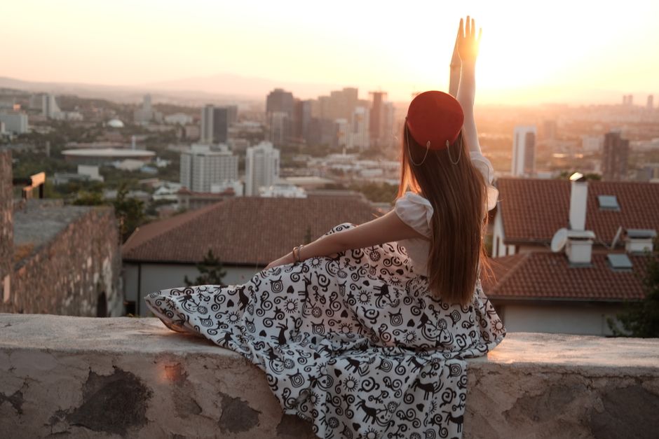 Silhouette of a woman seated on a wall, reaching towards a city skyline at sunset.