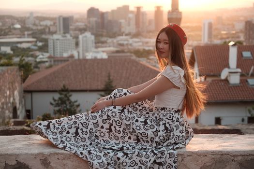 A woman sits on a rooftop at sunset with a cityscape backdrop, capturing urban tranquility and elegance.