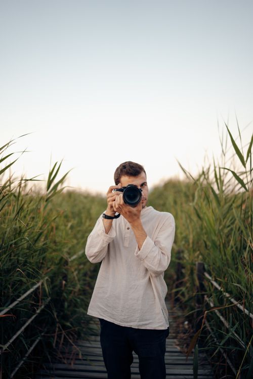 Free A young man photographing in a lush outdoor setting on a boardwalk during summer. Stock Photo