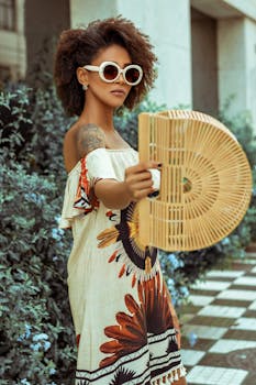 一位戴着太阳镜的时尚女性，在户外穿着时髦连衣裙，手拿柳条扇子