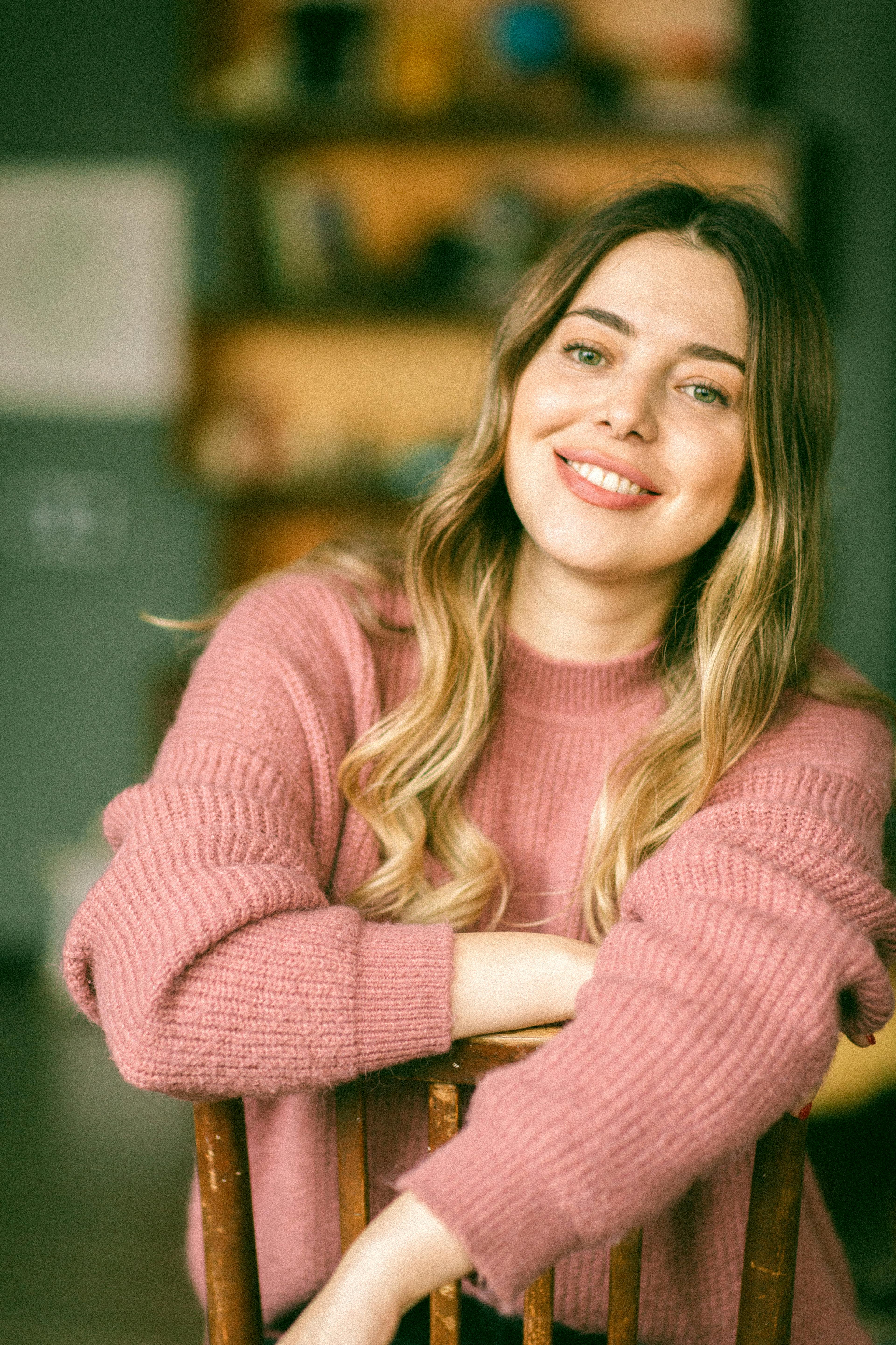 A young woman with long hair, smiling while leaning on a wooden chair in a cozy indoor setting.