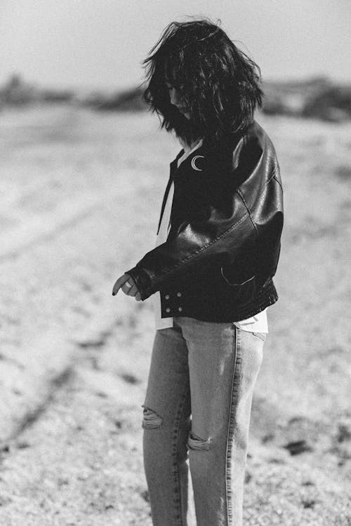 Brunette Woman in Leather Jacket and Ripped Jeans Standing on Sandy Beach