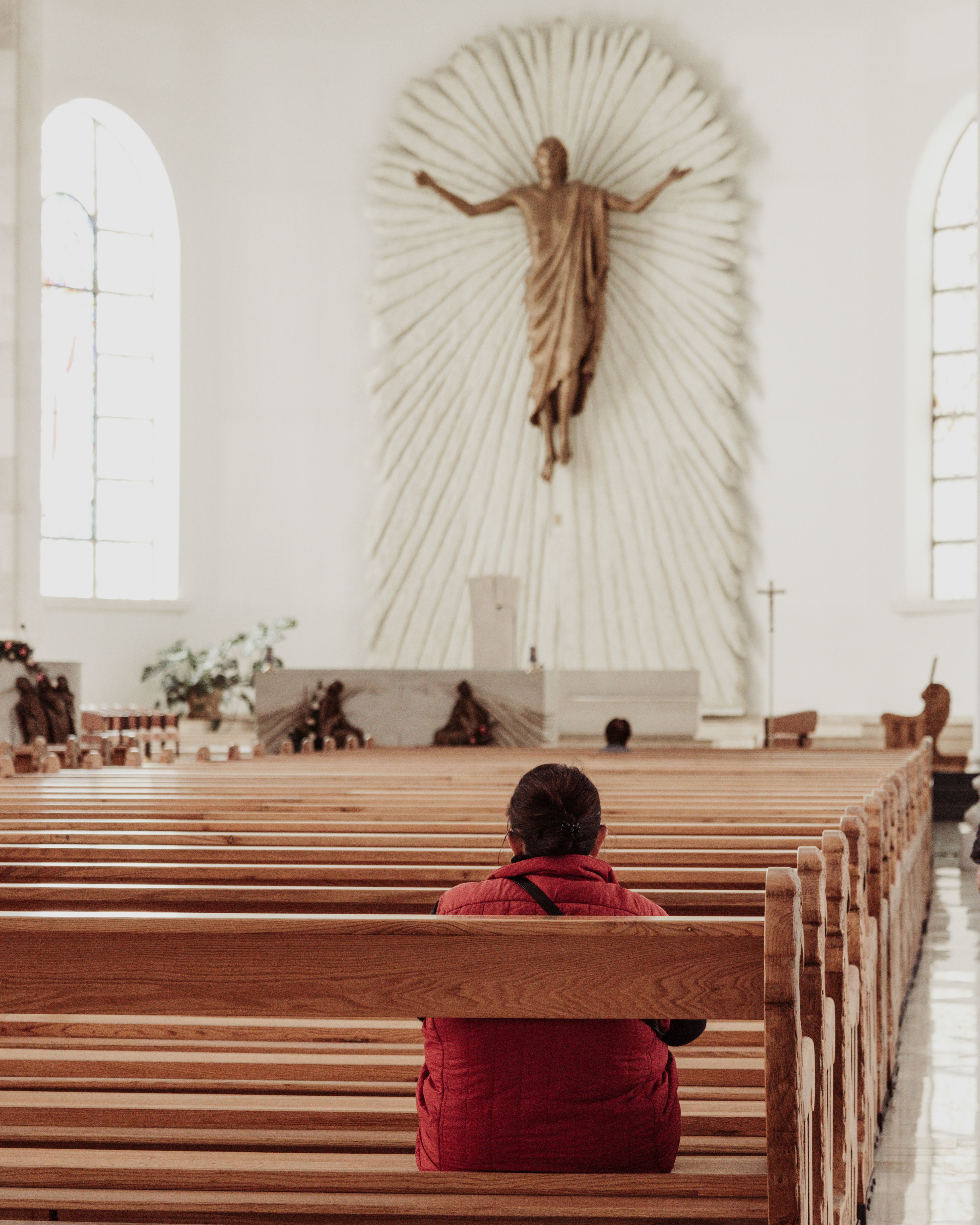 A woman in a red jacket sits alone, praying in an empty church, with a large sculpture in the background.