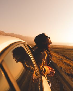 A serene moment as a woman leans out of a car window, enjoying a sunset on a rural road.