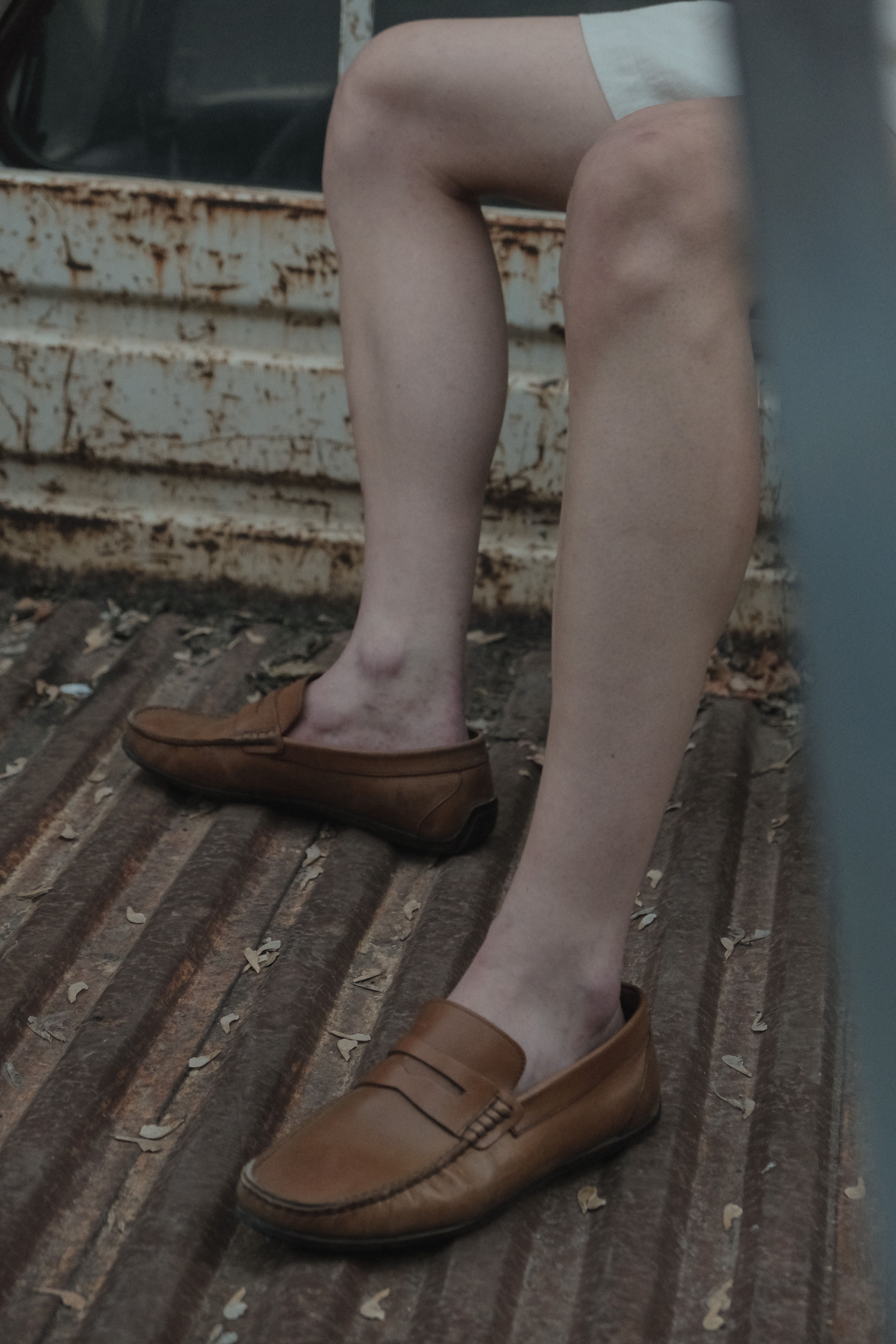 Close-up of legs wearing brown loafers against a rustic, urban backdrop.