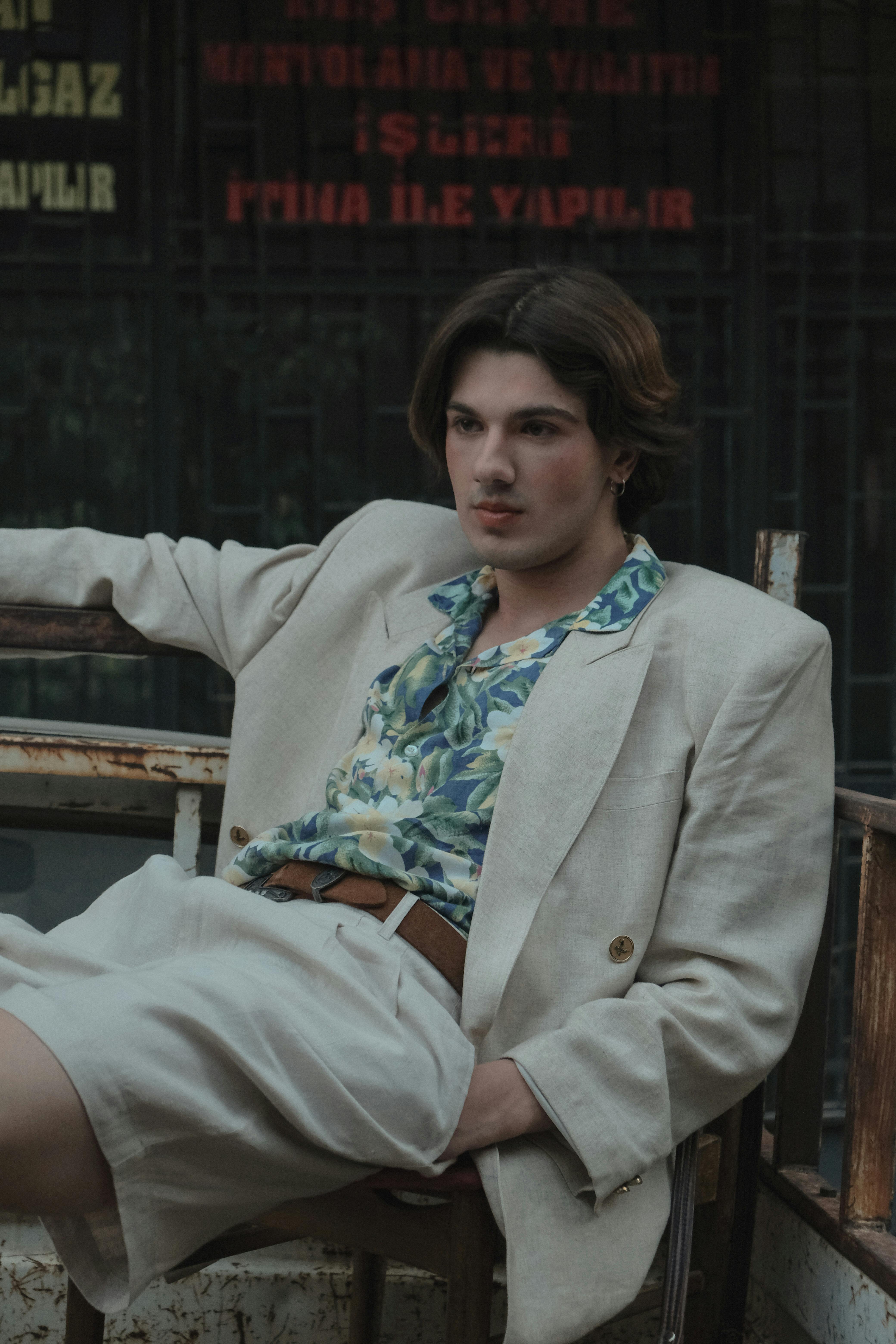 A fashionable young adult man in a casual outfit sitting confidently in an urban environment.