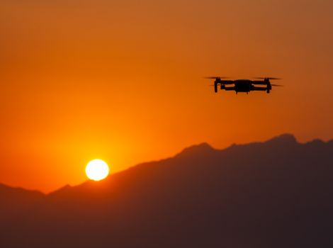 A silhouette of a drone flying against a stunning sunset over mountain peaks.