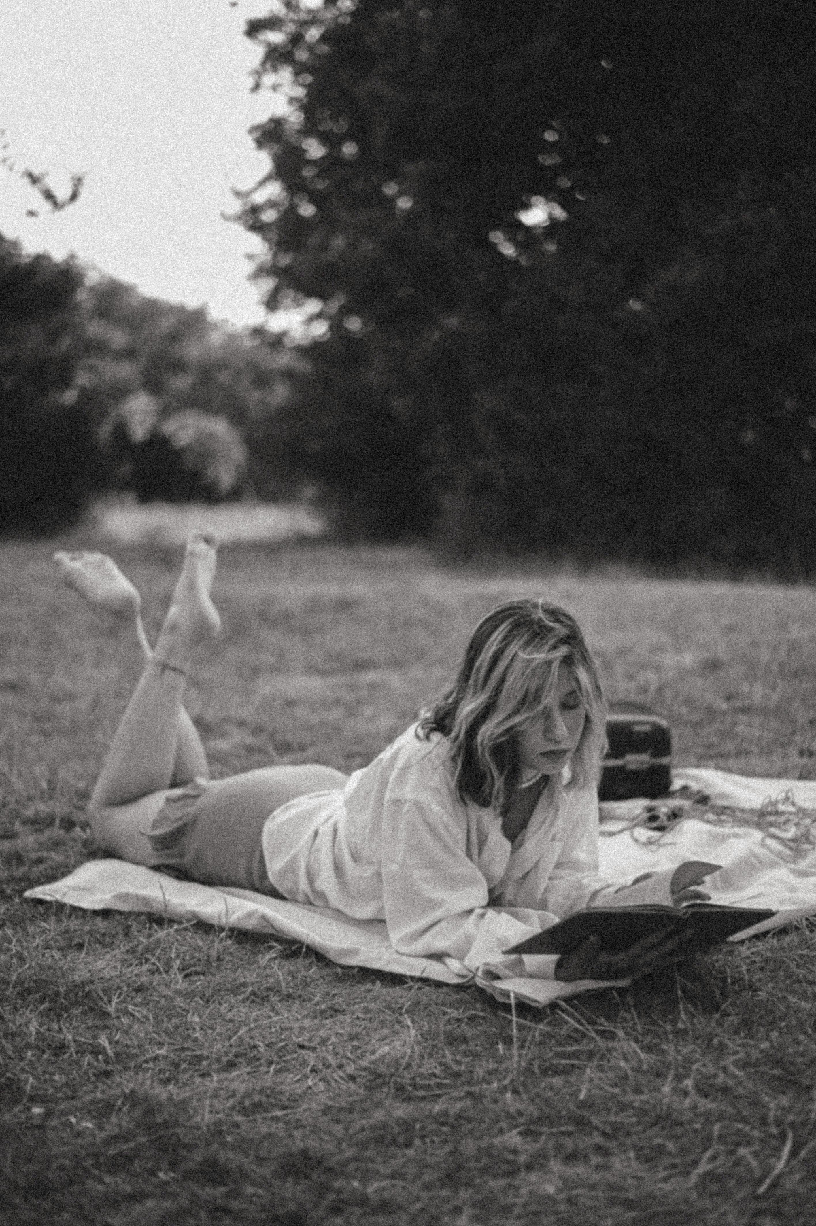 A young woman lies on a blanket outdoors, enjoying a summer afternoon reading a book.