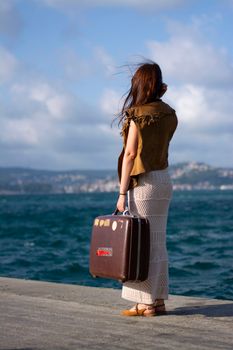 Woman standing by the sea with a suitcase on a breezy day, travel concept.