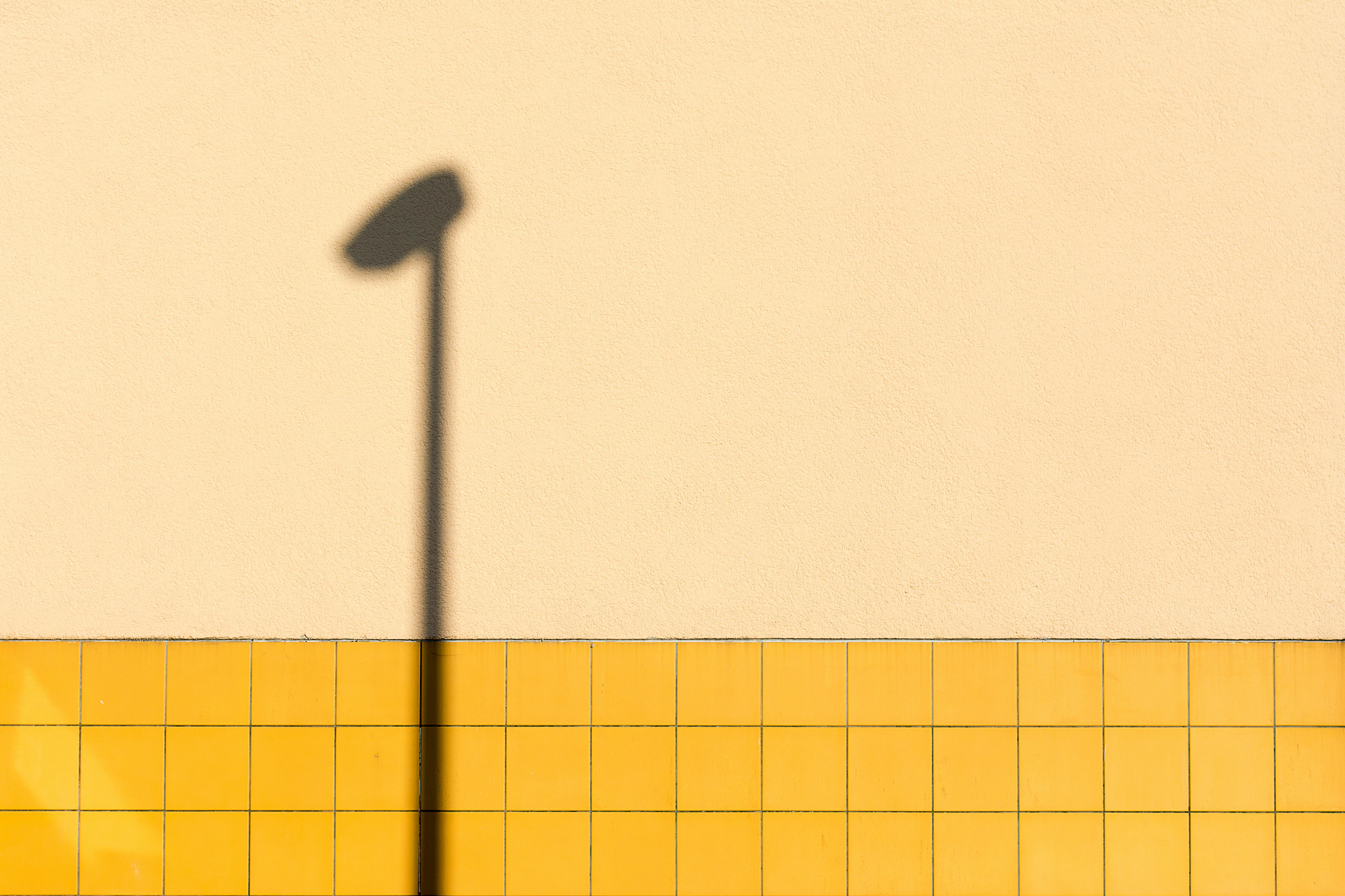 A minimalist image featuring the shadow of a streetlamp on a yellow tiled wall with a neutral background.