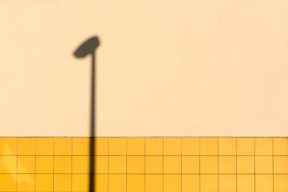 A minimalist image featuring the shadow of a streetlamp on a yellow tiled wall with a neutral background.