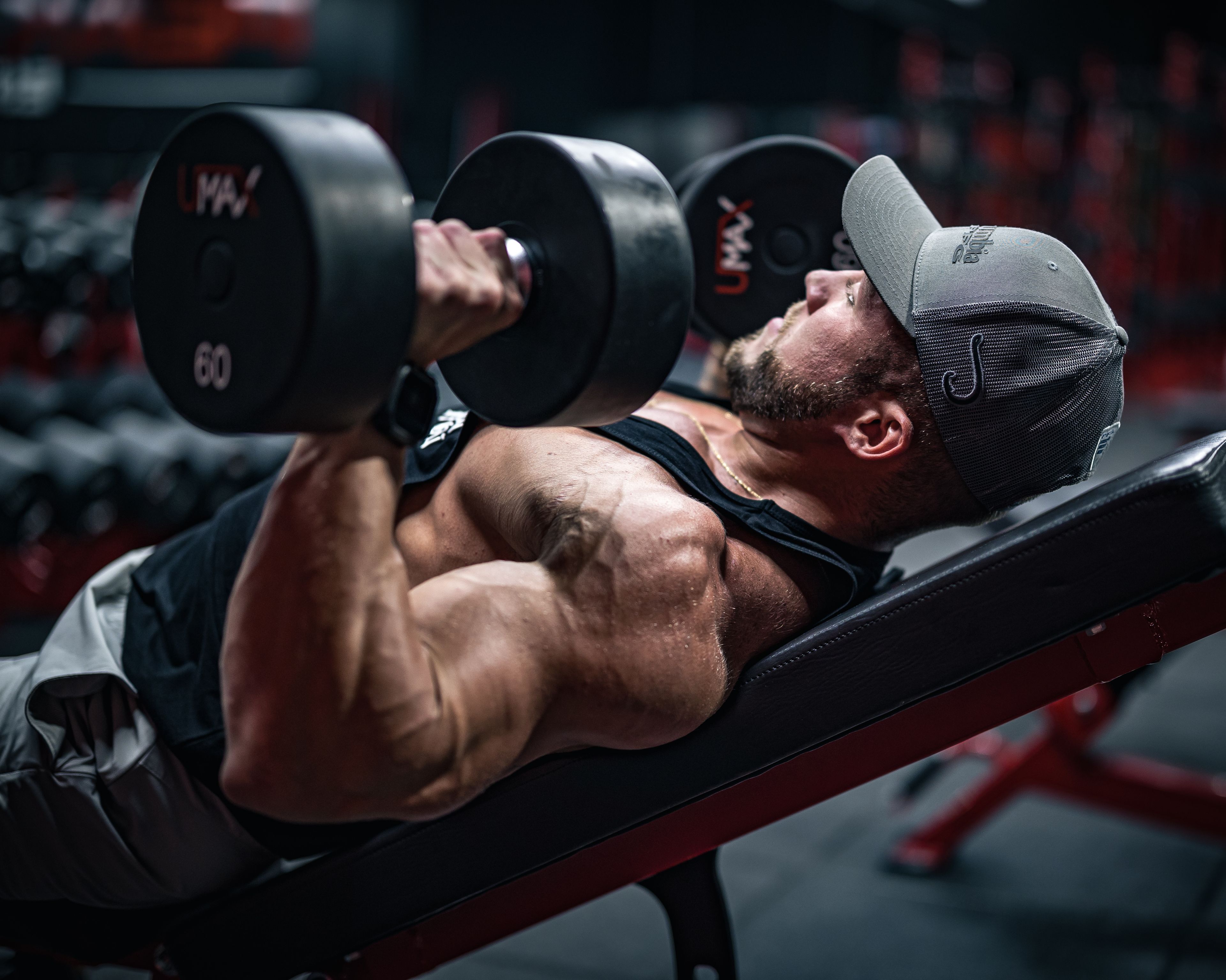 A muscular man focused on weightlifting exercises with dumbbells in a gym setting.