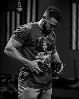 A muscular man adjusts his gym belt while standing indoors, portrayed in a dramatic black and white setting.