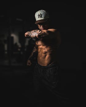 A muscular man with tattoos stretches in a dark gym environment, showcasing his physique.