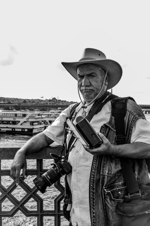 Free Black and white photo of a man with camera and hat on a bridge overlooking a river, exuding a travel vibe. Stock Photo