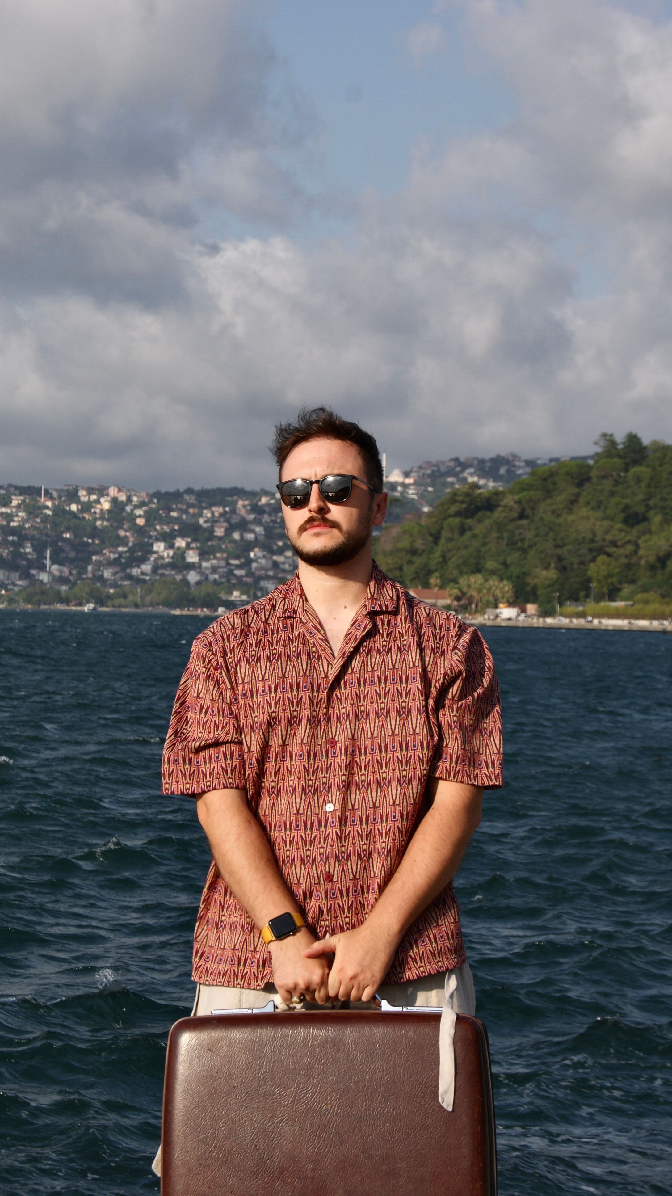 Man with sunglasses posing by Bosphorus in Istanbul, showcasing fashion and scenic backdrop.
