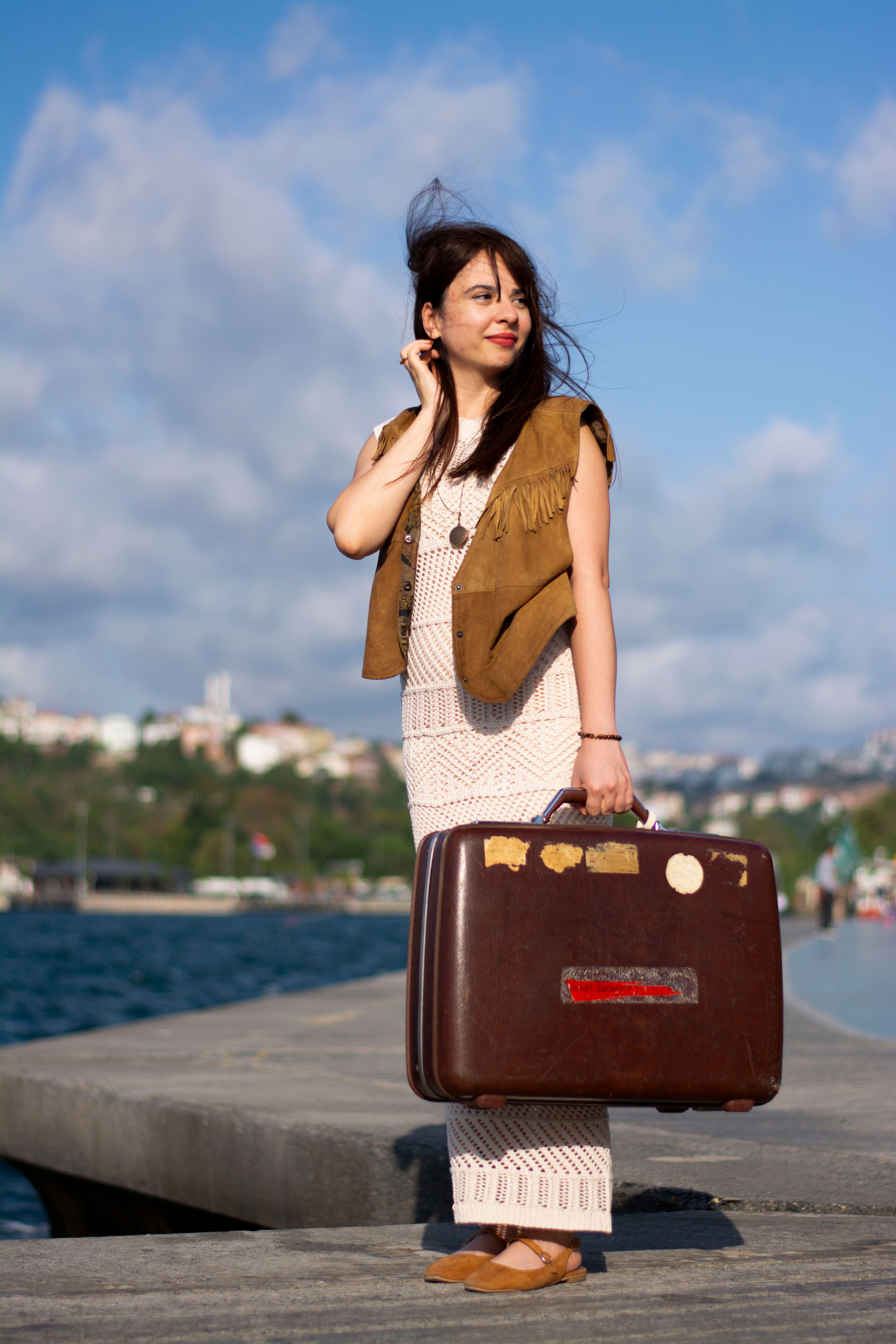 Woman in boho attire stands with vintage suitcase by the seashore, evoking travel and adventure.