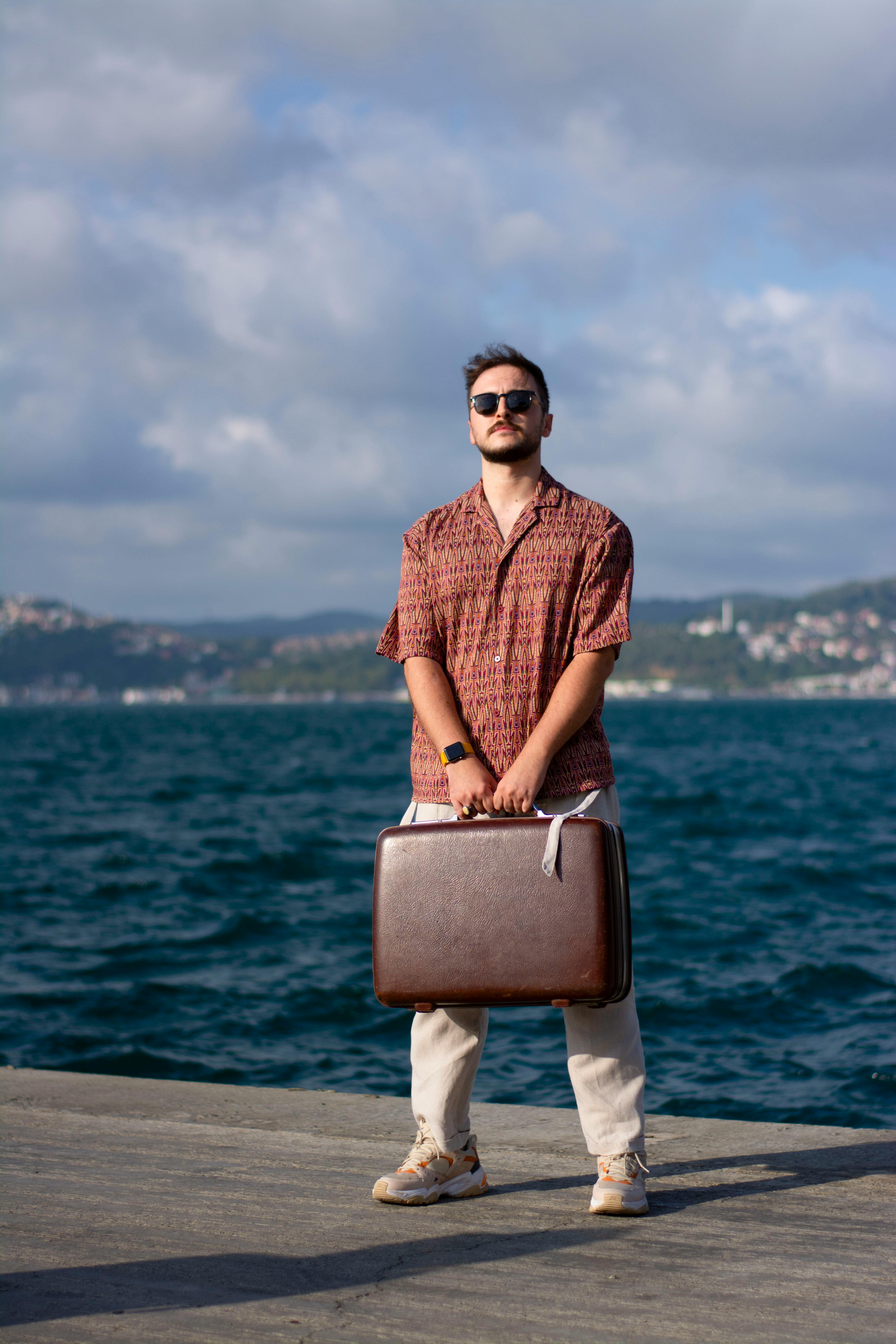 A fashionable man stands on a seaside pier holding a suitcase on a sunny day.