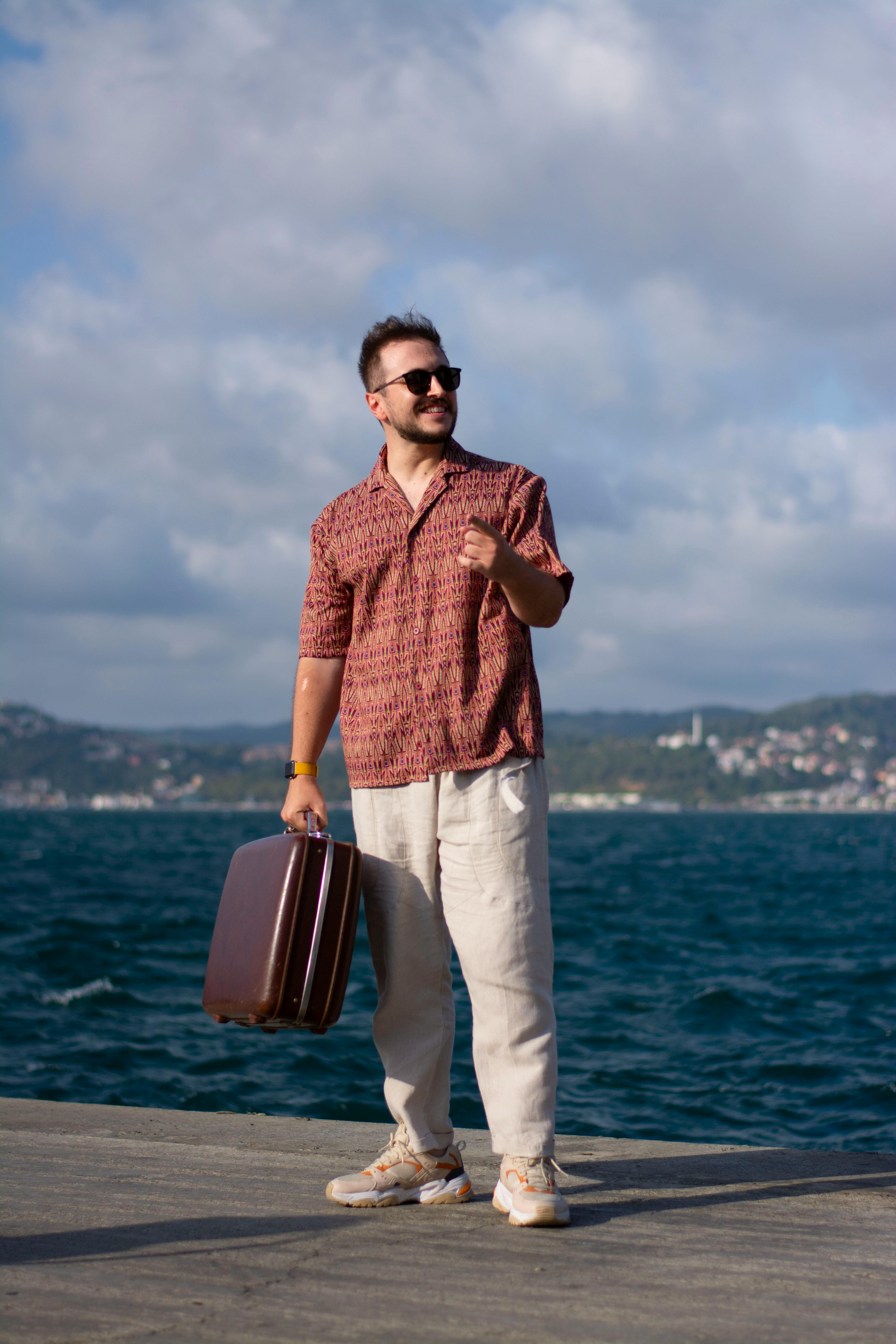 Smiling man in sunglasses holds suitcase by the sea, embracing vacation vibes.