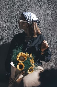 A woman in a headscarf and sunglasses holding sunflowers, captured in artistic light.