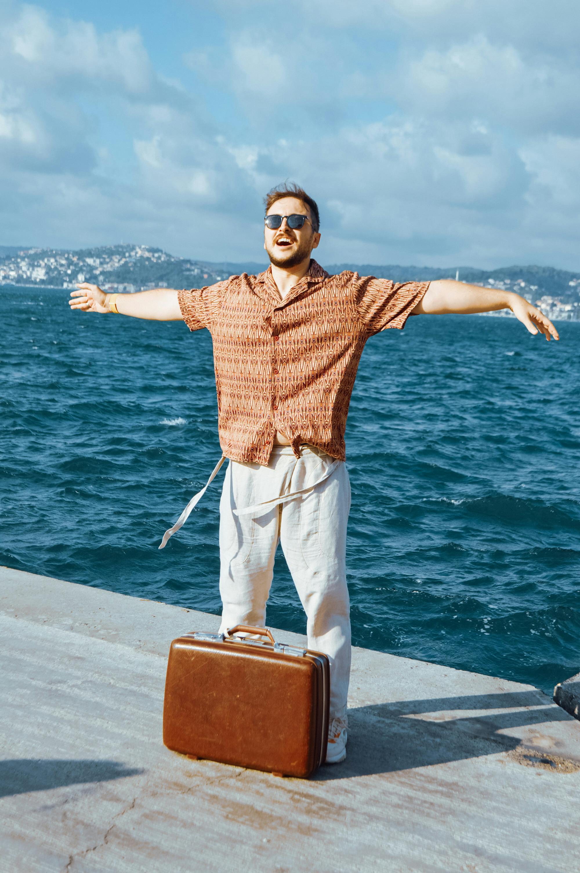 A man with sunglasses stands joyfully by the sea in Istanbul with arms wide open.