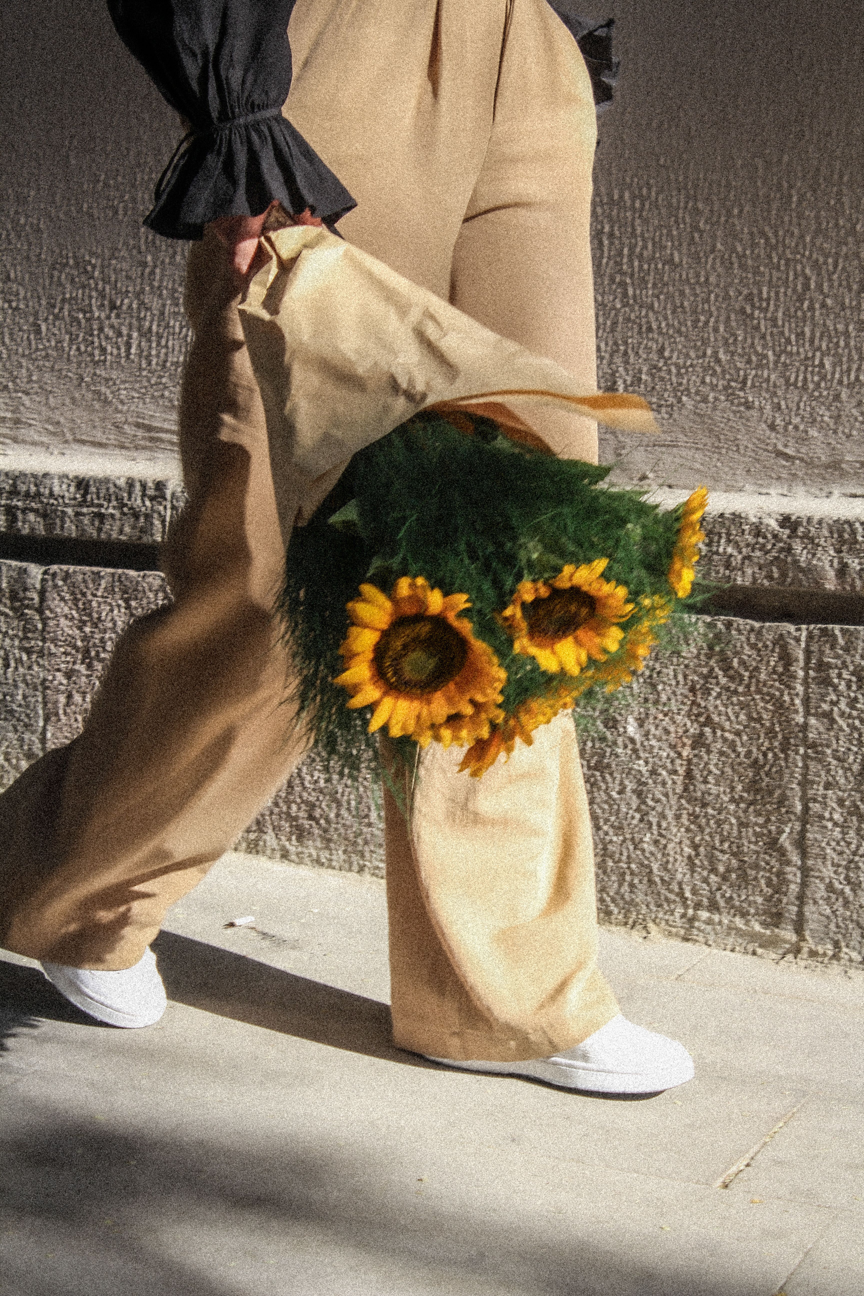 Stylish woman in beige pants holding a sunflower bouquet while walking on a sunlit sidewalk.
