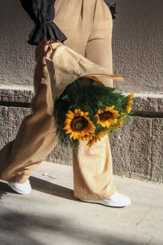Stylish woman in beige pants holding a sunflower bouquet while walking on a sunlit sidewalk.