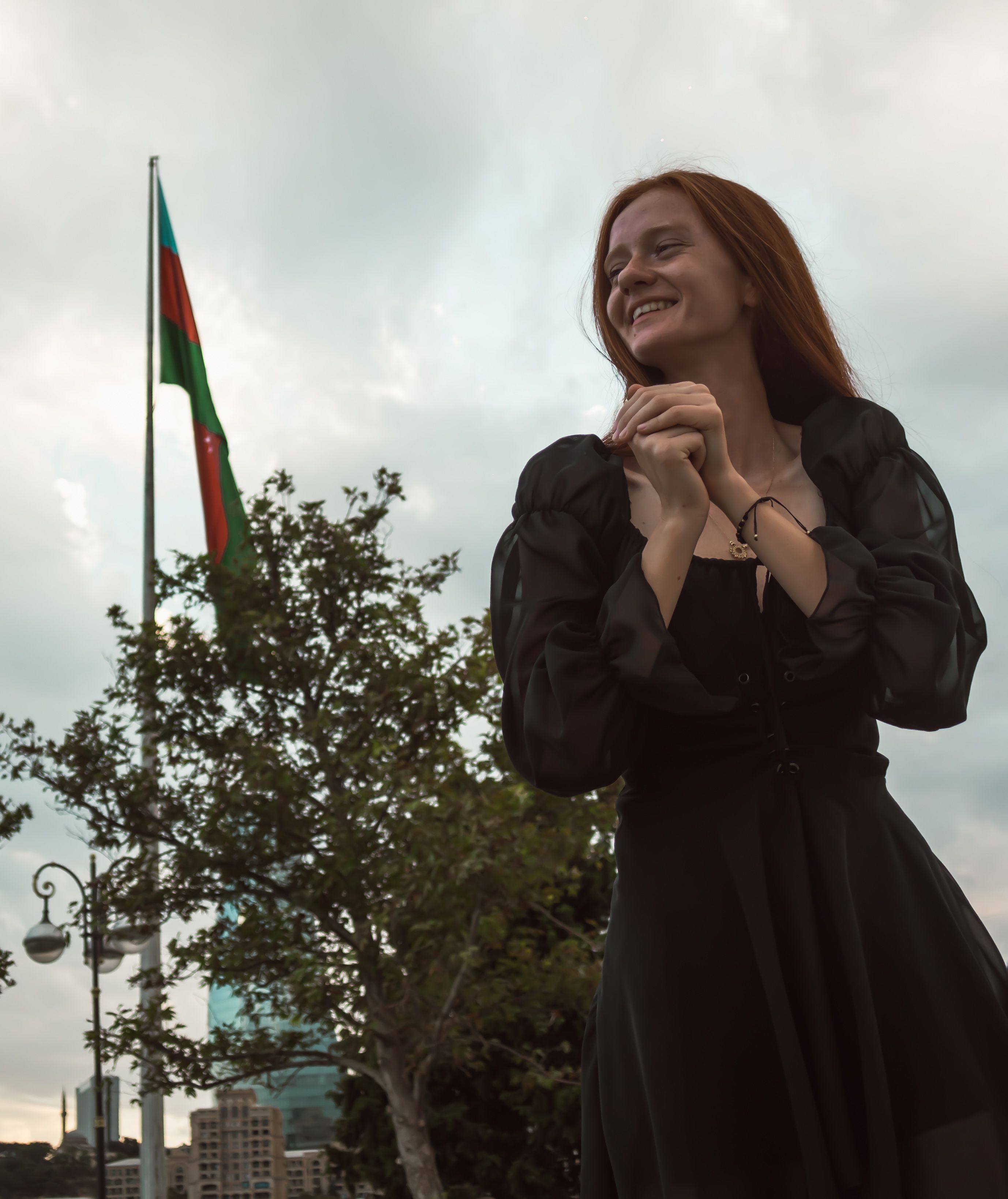 A redheaded woman in a black dress smiles with an Azerbaijani flag in the background in an outdoor setting.