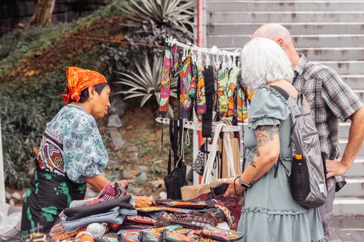 A local artisan selling colorful textiles to customers at a market stall outdoors.