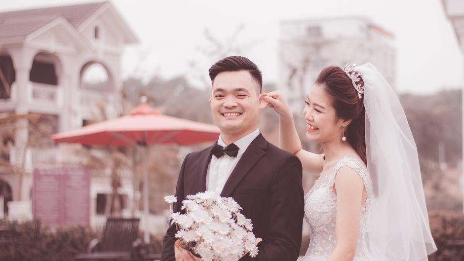 Smiling bride playfully adjusts groom's attire during outdoor wedding ceremony.