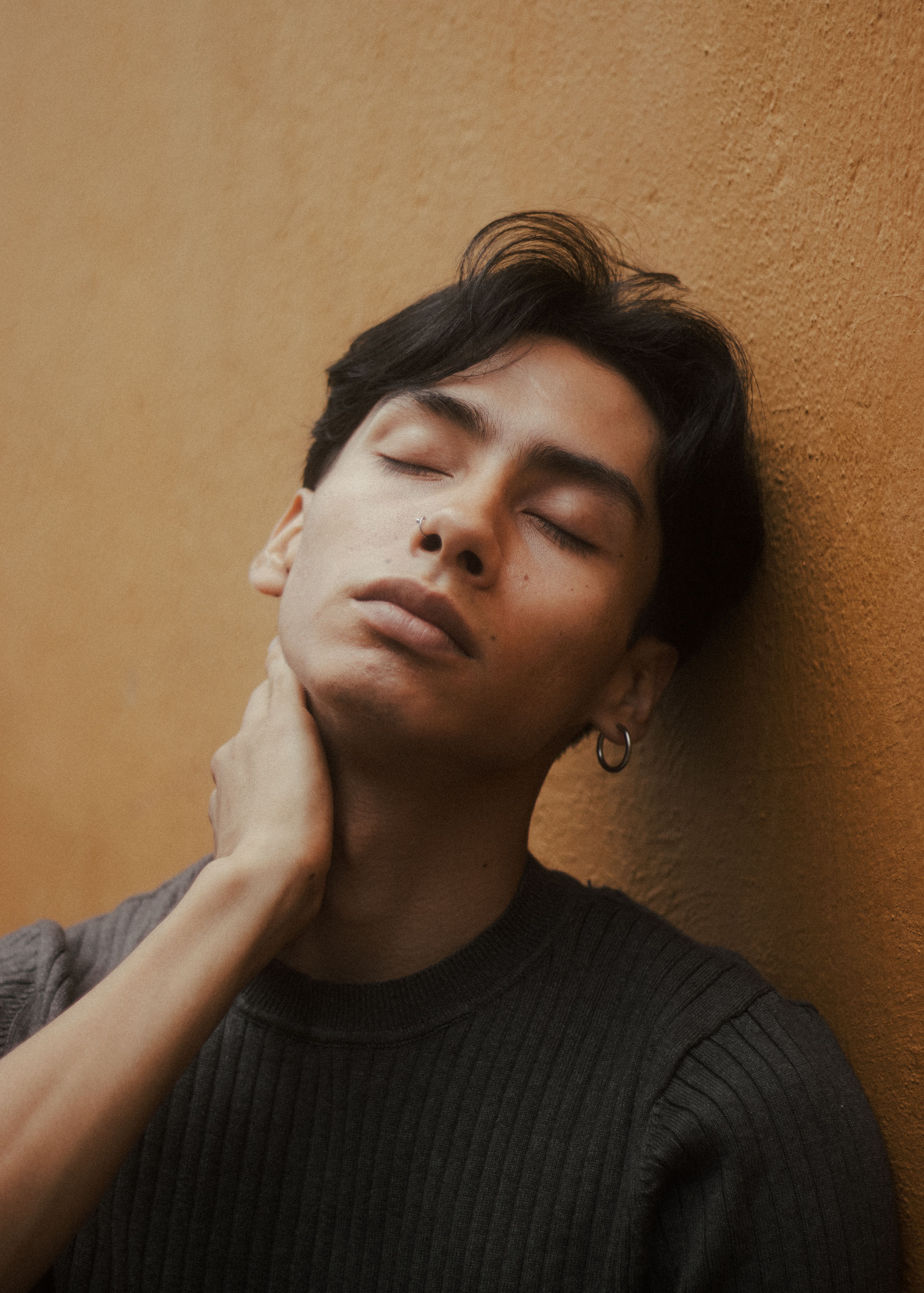 Close-up portrait of a young man with eyes closed, leaning against a wall, hand on neck.