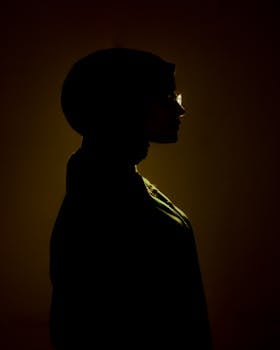 Artistic silhouette portrait of a woman with glasses wearing a hijab, against a warm background.