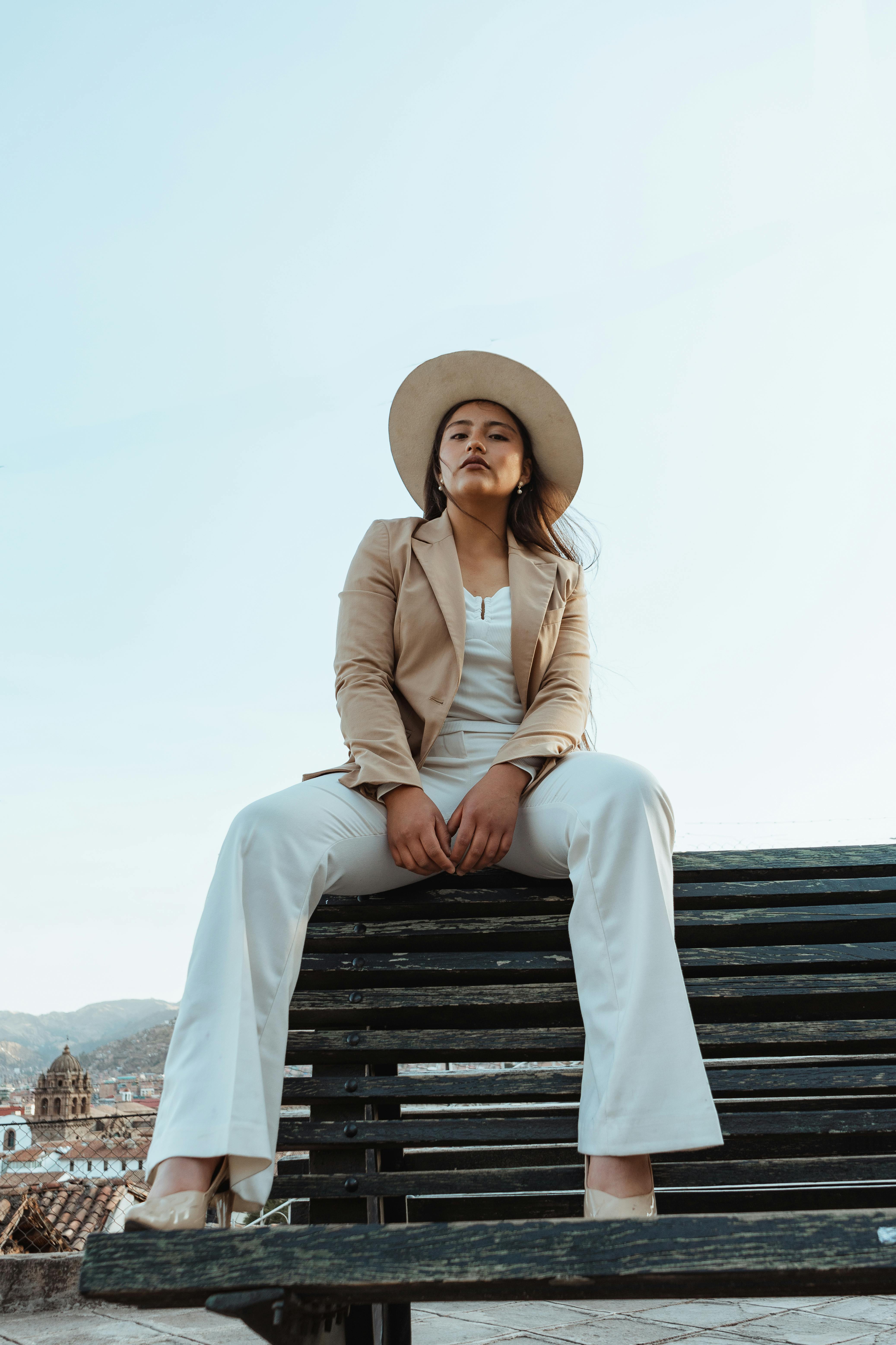 Stylish woman in elegant attire and hat posing on a bench against a clear sky in Cusco, Peru.