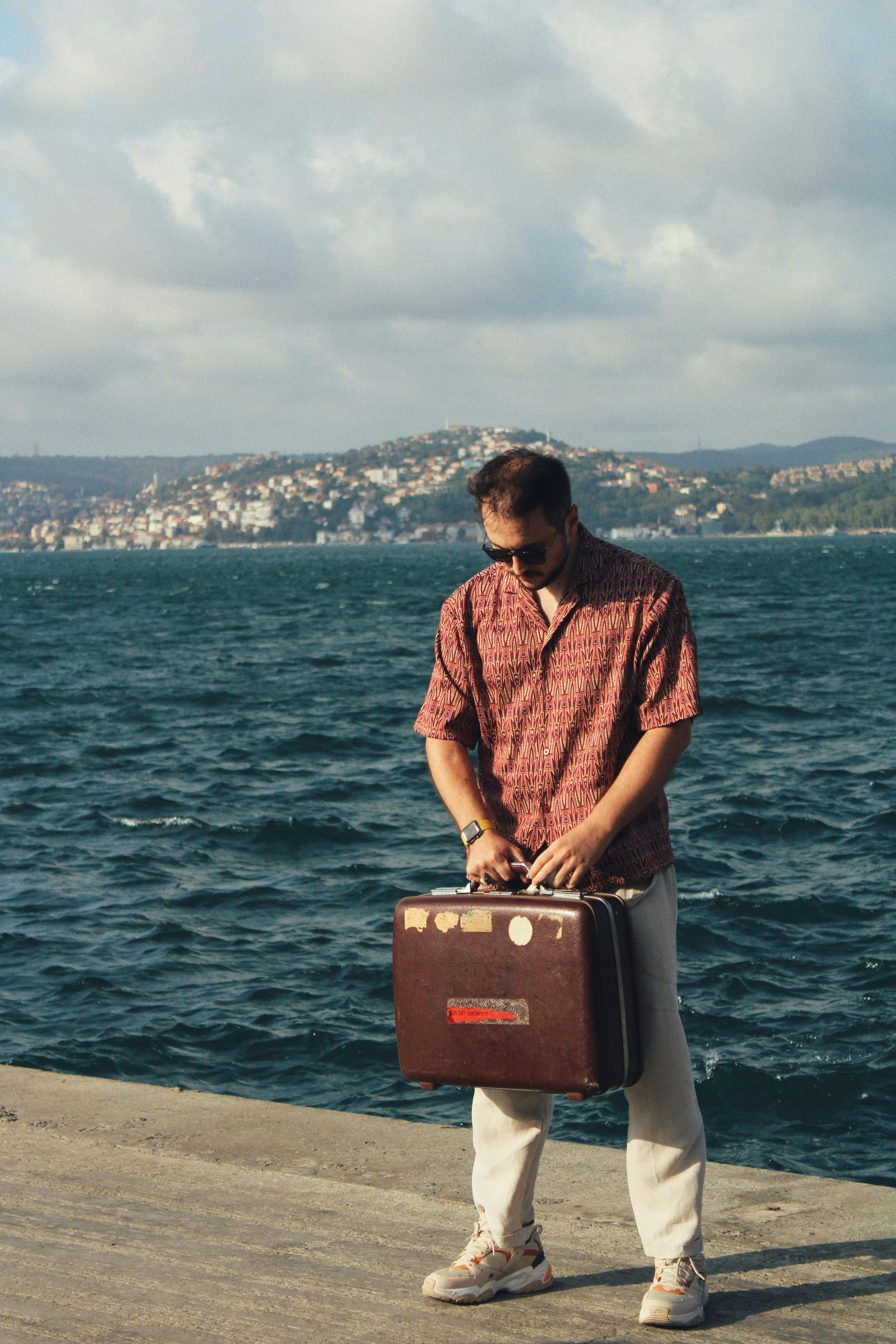 Man standing with a suitcase on a promenade by the sea under a cloudy sky.