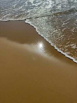 Close-up of ocean waves creating patterns on a sandy beach with sunlight reflection.